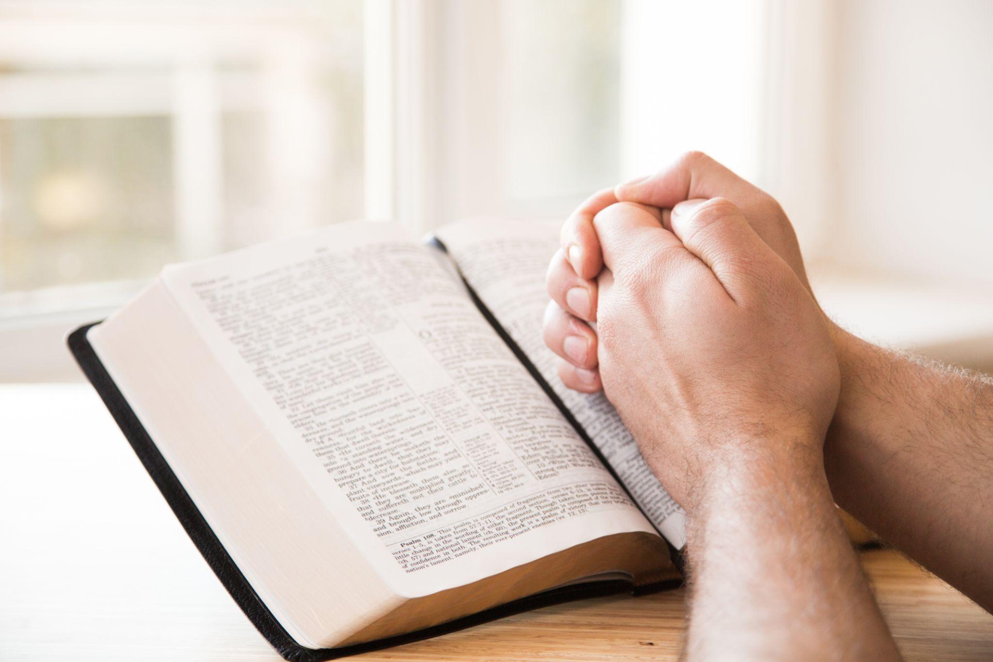 Hands folded in prayer resting on an open Holy Bible in bright, soft window light