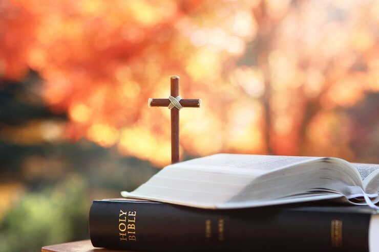A small wooden cross standing on an open Holy Bible, autumn leaves blurred behind